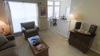 This single-story residence showcases an open-concept floor plan that seamlessly connects the living and dining areas, creating a cohesive and functional living environment. The living area is comfortably appointed with plush wall-to-wall carpeting in a neutral tone, providing a warm foundation for the space. A cushioned armchair and a wooden coffee table serve as focal points, complemented by a wooden chest of drawers and a floor lamp that adds warm ambient lighting.

Natural light is a defining characteristic of this home, with multiple large windows dressed in white horizontal blinds allowing ample daylight to fill the interior. The dining area, visible from the living space, features a small round dining table with chairs and is further brightened by a bay window arrangement, enhancing the open and airy feel of the home.

A partial wall with a countertop pass-through separates the dining area from what appears to be the kitchen, maintaining visual connectivity while defining each functional zone. Neutral-toned walls throughout lend a clean, versatile backdrop, and the overall design reflects a practical yet comfortable living environment suited for everyday living.