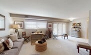 This single-floor residence offers a thoughtfully arranged layout encompassing two bedrooms, a bathroom, a galley-style kitchen, and an open living and dining area. The living space is generously sized, featuring soft wall-to-wall carpeting, neutral tones, and multiple windows that allow natural light to fill the room. A set of French doors provides direct outdoor access, enhancing the sense of openness. The combined dining area sits adjacent to the living space, making it well-suited for everyday living and entertaining. The galley kitchen is efficiently designed with warm oak cabinetry lining both sides, laminate countertops, tile flooring, and white appliances including a gas range and refrigerator, providing ample storage and counter space. The primary bedroom is a comfortable retreat featuring carpeted flooring, a large window dressed with plaid curtains, and a soft, neutral color palette. The second bedroom is equally inviting, with carpeted floors, a window with natural light, and a cozy, relaxed atmosphere. The bathroom is cleanly appointed with white tile wainscoting, a vanity with under-cabinet storage, and a shower-tub combination. Throughout the residence, a consistent use of warm, neutral tones and eclectic rustic décor accents creates a cohesive and welcoming ambiance.