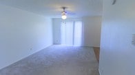 This single-floor residential space offers a clean and functional layout designed for comfortable everyday living. The main living area is a standout feature, showcasing wall-to-wall plush carpeting that adds warmth and comfort underfoot. A ceiling fan with a built-in light fixture is centrally mounted on the textured ceiling, providing both air circulation and ambient lighting throughout the room. A large sliding glass door dressed with vertical blinds serves as a prominent architectural element, allowing an abundance of natural light to fill the space while offering privacy and light control as needed. The room's neutral, white-toned walls create a bright and airy atmosphere, lending a sense of openness and versatility for a variety of interior design styles. A transition to hard flooring is visible at the edge of the living area, suggesting a defined boundary leading to an adjacent space such as a dining area or hallway. The overall ambiance of the space is bright, clean, and move-in ready, making it well-suited for those seeking a straightforward and welcoming home environment.