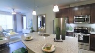 This single-floor residence showcases a thoughtfully designed open-concept layout that seamlessly connects the kitchen and living areas. The kitchen is a standout feature, boasting a generously sized curved island topped with speckled granite countertops that provide ample prep and dining space. Rich dark-wood cabinetry lines the walls, offering abundant storage, while stainless steel appliances, including a French door refrigerator, lend a sleek, modern aesthetic. A stainless steel undermount sink sits centrally in the island beneath two stylish pendant lights that cast warm, focused illumination over the workspace. The neutral green-toned walls create a calming and cohesive palette throughout, complementing both the cabinetry and decorative accents. A large mirror in the adjoining space enhances the sense of depth and openness, while natural light filters in through multiple windows, brightening the living area beyond. The living space is visible from the kitchen, reinforcing the open and connected feel of the floor plan. Overall, the residence combines functional design with contemporary finishes, offering a comfortable and visually appealing living environment suited for both everyday living and entertaining.