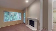 This well-appointed single-floor space offers a warm and inviting atmosphere highlighted by thoughtful architectural details. The living area serves as the centerpiece of the home, drawing the eye upward with soaring vaulted ceilings that lend an impressive sense of openness and volume. A classic white brick fireplace anchors one wall, complete with a clean white mantel and a raised hearth, creating a cozy focal point that adds both character and charm to the room. Soft, neutral-toned carpeting extends throughout, providing warmth underfoot and complementing the light, beige wall palette. Natural light fills the space generously through a large window fitted with horizontal blinds, offering a pleasant view of mature trees and lush green surroundings. A glass-panel door adjacent to the window provides convenient access to the outdoors while further enhancing the connection to the natural environment outside. Recessed ceiling lighting ensures the space remains bright and well-illuminated at any time of day. The overall ambiance is calm and comfortable, with a neutral color scheme and clean finishes that create a versatile backdrop suited to a variety of interior styles.