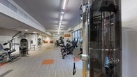 This single-floor fitness center offers a thoughtfully designed space built to support a comprehensive workout routine. The layout is expansive and open, with a long floor plan that allows for generous spacing between equipment, promoting ease of movement and a comfortable exercise environment.

A row of cardio machines, including stationary bikes, ellipticals, and stair climbers, lines one side of the room, each unit equipped with individual digital displays for tracking performance metrics. On the opposite side and toward the rear, strength training stations and cable machines provide options for resistance-based workouts.

The flooring features a patterned carpet with orange accent squares that define specific workout zones, adding both visual interest and functional organization to the space. Overhead, linear fluorescent lighting runs the length of the ceiling, ensuring consistent, bright illumination throughout. The ceiling height contributes to an airy, open feel. Large windows at the far end allow natural light to filter in, enhancing the overall ambiance. The clean, neutral color palette of white walls and light gray tones throughout creates a focused and energizing atmosphere suited for fitness activities.