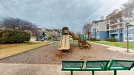 This community outdoor space serves as a central recreational hub for residents, offering a variety of amenities designed for leisure and social interaction. The playground is a standout feature, equipped with a multi-lane slide, a tunnel climber, and green steel play structures set atop a wood chip safety surface, providing a safe and engaging environment for children. Adjacent to the playground, green metal benches offer convenient seating for supervising adults. A covered octagonal pavilion anchors another portion of the space, featuring a warm wooden ceiling supported by sturdy metal posts. Inside the pavilion, green metal picnic tables provide shaded seating, making it ideal for gatherings and outdoor dining. A standalone charcoal grill is positioned nearby, adding to the area's outdoor entertaining functionality. Concrete walkways connect the various amenities throughout the space, and a bike rack is available for resident use. The grounds are landscaped with open grassy areas, mature trees, and decorative shrubs that add color and greenery to the environment. Multi-story residential buildings border the space on multiple sides, giving the area an intimate, community-centered feel.