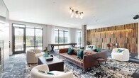 This expansive single-floor amenity lounge is a masterfully designed social space that seamlessly blends industrial character with contemporary elegance. The open layout is anchored by a tufted caramel leather sofa and coordinating seating arrangements, all set atop richly patterned area rugs that add depth and texture to the space.

A dramatic gray tile feature wall serves as the focal point of one end of the lounge, housing a sleek linear fireplace with a striking green geometric tile surround and a wall-mounted television above. Adjacent to this, a series of exposed brick archways frame intimate banquette-style seating areas with warm gold accents, adding a timeless architectural detail that contrasts beautifully with the modern finishes throughout.

On the opposite end, floor-to-ceiling windows flood the space with natural light and offer open views of the surrounding landscape, with a shuffleboard table and a reclaimed wood accent wall adding recreational and visual interest. A full-service wet bar area features a bold chevron-patterned tile backsplash, dark cabinetry, a marble-topped island, and stainless steel appliances beneath a warm wood-planked ceiling.

Sputnik-style chandeliers, lush indoor plants, and curated decor complete the sophisticated yet inviting atmosphere of this thoughtfully designed communal space.
