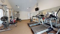 Located at 8799 Old Colony Way in Alexandria, Virginia, this single-floor fitness center is a well-equipped exercise facility designed to support a comprehensive workout routine. The space features vaulted ceilings that lend an open, airy feel, while neutral-toned walls and carpeted flooring create a clean, functional environment throughout.

The cardio zone is outfitted with multiple treadmills, a stationary bike, and an elliptical trainer, providing a variety of aerobic exercise options. A wall-mounted flat-screen television is positioned for easy viewing during workouts. Large wall mirrors span one side of the room, enhancing the sense of space and allowing users to monitor their form.

The strength training area includes a multi-functional cable and Smith machine by Life Fitness, an adjustable weight bench, a dumbbell rack with a full range of free weights, and additional resistance equipment. Natural light filters in through several windows fitted with blinds, offering pleasant views of the surrounding greenery.

The facility also includes a small storage unit and a dedicated exit door, ensuring both convenience and safety. The overall layout is thoughtfully organized, maximizing usable floor space while keeping equipment accessible and the environment comfortable for exercisers of all levels.