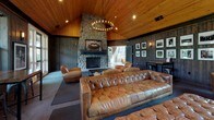 This single-floor lounge space blends rustic charm with modern comfort through a cohesive and carefully considered design. The defining architectural feature is the soaring vaulted ceiling clad in warm natural wood planks, which lends the space an open, airy quality and a distinct lodge-like character. A large circular chandelier anchors the ceiling, complementing the recessed lighting throughout.

At one end of the room, a commanding floor-to-ceiling stone fireplace serves as the focal point, constructed from rounded river rock and topped with a wide mantel. Framed artwork and photographs are displayed across the dark wood-paneled walls, adding a curated, personal touch to the interior.

The seating arrangement is generous and inviting, centered around a substantial tufted leather Chesterfield sofa paired with several cognac leather swivel club chairs. A low bench and area rugs define the gathering zones, creating distinct areas for conversation and viewing. Large wall-mounted flat-screen televisions are positioned for optimal viewing from multiple seating areas.

Natural light filters in through multiple windows along the walls, offering views of the surrounding outdoor greenery. French doors provide direct access to the exterior. A high-top table with bar stools occupies a corner, adding a casual dining or social option. The overall ambiance is warm, relaxed, and richly appointed — suited for both casual lounging and social gatherings.