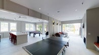 This single-floor recreation space is designed for leisure and entertainment, combining a clean, contemporary aesthetic with a functional layout. The room is finished with light-tiled flooring and smooth white ceilings fitted with recessed lighting, creating a bright and open atmosphere throughout.

At the heart of the space, a full-size ping pong table takes center stage, while a sleek pool table anchors the adjacent area, making this an ideal setting for social gatherings and friendly competition. Wall-mounted sconce lighting adds a modern accent along the grey feature wall, complementing the neutral color palette of the room.

A comfortable lounge seating area is positioned near large sliding glass doors that open to an outdoor area, inviting abundant natural light and providing easy indoor-outdoor flow. Additional windows along the perimeter further enhance the sense of openness and connection to the surrounding greenery.

Ceiling fans are visible in the background, ensuring comfort across the space, while the thoughtful separation of activity zones — gaming, lounging, and transition areas — gives the room a well-organized yet relaxed feel. Overall, this recreation space delivers a polished, welcoming environment suited for community or shared-use amenity purposes.