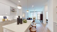 This well-appointed single-floor residence showcases a clean, contemporary design throughout. The open-concept layout connects the kitchen and living area seamlessly, creating a bright and inviting communal space. The kitchen is a standout feature, outfitted with high-gloss white upper cabinetry, light wood lower cabinetry, stainless steel appliances including a refrigerator, range, and microwave, and a generous white quartz island with bar seating illuminated by modern pendant lighting. Light wood flooring flows throughout the space, adding warmth to the modern aesthetic.

The living room is bathed in natural light from floor-to-ceiling windows and a glass door that opens to a private balcony, offering outdoor views. A ceiling fan overhead adds comfort to the space.

Two bedrooms provide comfortable retreats, each featuring large windows that invite natural light. The primary bedroom is spacious and includes direct access to an en-suite bathroom, which is equipped with dual sinks set in a quartz vanity, gold fixtures, a walk-in tiled shower, and a walk-in closet. The second bathroom offers a tiled tub-shower combination, a single vanity, and crisp, modern finishes consistent with the rest of the home.