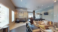 The residents lounge is a thoughtfully designed communal space that seamlessly blends relaxation, recreation, and productivity into one welcoming environment. The open floor plan creates a generous sense of space, with distinct zones that cater to a variety of activities and social settings.

At the heart of the lounge, a full-size billiards table takes center stage, illuminated by a striking cluster of pendant lights with gold-toned fixtures that add a warm, sophisticated glow. Surrounding it, large sectional sofas dressed in neutral tones and accented with decorative pillows offer comfortable seating for casual gatherings.

A dedicated workspace area toward the back of the lounge provides computer stations and desk seating, making it a practical retreat for residents who need a quiet place to work. Nearby, a stone-accented column adds a natural architectural element that anchors the space.

Smaller bistro-style tables with chairs line one wall, ideal for intimate conversations or solo use. Wall sconces and recessed lighting throughout ensure the space is well-lit, while neutral-toned walls and tile flooring with varied textures contribute to the lounge's polished, contemporary ambiance.