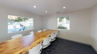This Conference Center offers a well-appointed, single-floor meeting space designed to foster productivity and collaboration. Entry is through a sleek glass-paneled door etched with "Conference Center," opening into a bright, welcoming corridor that leads directly into the main meeting room.

The centerpiece of the space is a remarkable live-edge wood conference table, showcasing the natural grain and organic contours of the slab. Surrounded by modern white rolling chairs, the table comfortably accommodates a sizeable group, making it well-suited for team meetings, presentations, or client discussions.

The room is finished with neutral gray walls and dark carpeted flooring, creating a clean, professional aesthetic. Recessed lighting provides even, consistent illumination throughout. Multiple horizontal windows along the walls invite an abundance of natural light and offer views of the surrounding greenery and outdoor environment, lending the space an open, airy feel.

A large-scale contemporary cityscape painting adorns one wall, adding a vibrant artistic touch that complements the room's modern sensibility. An interior window connects the conference room to an adjacent hallway or waiting area, enhancing spatial connectivity within the facility. Overall, the Conference Center balances functional design with distinctive character, providing an inspiring environment for professional use.