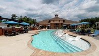 This community amenity space combines an expansive outdoor pool area with a fully equipped interior clubhouse. The centerpiece is a generously sized freeform pool with crystal-clear blue water, featuring built-in in-water lounge chairs for relaxed sunbathing and easy entry steps. The surrounding pool deck is finished with decorative pavers and outfitted with numerous chaise lounges shaded by vibrant blue market umbrellas, providing ample space for outdoor recreation and relaxation. A charming clubhouse building with a distinctive pitched roof and red-and-brick exterior anchors the backdrop of the pool area, with mature trees framing the perimeter. Inside, the clubhouse offers a thoughtfully designed interior with light wood-look tile flooring, recessed lighting, and a textured stone accent wall. A comfortable lounge area with blue upholstered seating and individual work desks creates a welcoming reception space. Glass-walled private offices or meeting rooms provide transparency while maintaining separation. A dedicated conference room with a full table and rolling chairs is ideal for group gatherings. A game room with a billiard table and large windows adds a recreational element, while a beverage and coffee station ensures convenience for all residents.