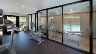 This fitness center is a bright, single-floor facility designed to support a well-rounded workout routine. The space is thoughtfully laid out with a selection of cardio and strength-training equipment, including a treadmill, elliptical, recumbent exercise bike, and a multi-function cable machine, ensuring users have access to a comprehensive range of exercise options.

One of the standout architectural features is the expansive floor-to-ceiling glass walls and large windows that line the space, flooding the interior with abundant natural light. These windows offer direct views of an outdoor swimming pool and mature, lush landscaping, creating an inviting and motivating atmosphere during workouts. Adjustable blinds provide the option for privacy when needed.

The floor is finished with durable dark rubber flooring, well-suited for fitness use, while recessed lighting in the ceiling supplements the natural light. A wall-mounted fan ensures comfortable air circulation throughout the space. The entrance features a glass door that opens directly to the outdoor grounds, reinforcing the seamless connection between the interior fitness area and the exterior environment. A welcoming fitness center mat marks the entry, and a small storage basket keeps accessories organized near the door. The overall ambiance is clean, functional, and open — ideal for daily exercise.