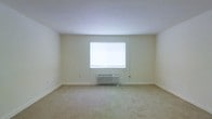 This single-floor space offers a straightforward, functional layout with a clean and understated aesthetic. The main room is finished with wall-to-wall carpeting in a neutral beige tone, providing a soft underfoot surface that contributes to the room's quiet, subdued atmosphere. The walls are painted in a light, off-white color, creating a bright and open feel throughout the space. A centrally positioned window allows natural light to filter into the room, enhancing the airy quality of the interior. Beneath the window, a wall-mounted HVAC unit provides both heating and cooling, ensuring year-round climate control. The room features standard ceiling height with smooth white ceilings and clean white baseboards that frame the perimeter of the space. The overall design is simple and unadorned, making it a highly adaptable environment well-suited for a variety of uses. The neutral palette and minimalist finishes throughout provide a blank canvas, allowing occupants to personalize and furnish the space according to their own needs and preferences.