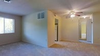 This single-floor residence offers a straightforward and functional layout designed for comfortable everyday living. The main living area is finished with wall-to-wall carpeting that adds a sense of warmth underfoot, while light-toned walls create a clean, neutral backdrop throughout the space. A ceiling fan with an integrated light fixture is centrally positioned overhead, providing both air circulation and ambient lighting to the room. A wall-mounted HVAC vent ensures climate control throughout the living area. The space transitions seamlessly into the kitchen, which is partially visible through a connecting doorway. The kitchen is equipped with essential appliances, including a full-sized refrigerator, making it a practical and ready-to-use cooking space. The open sightlines between the living area and kitchen promote a sense of connectivity and ease of movement. The overall design of this residence is practical and uncluttered, offering a blank canvas that is well-suited for personalization. The neutral color palette and simple finishes throughout make it an accessible and adaptable space for a variety of living arrangements.