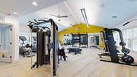 Located at Rolling Hills Circle in Easley, South Carolina, this fitness center delivers a comprehensive workout facility across a generously sized single-floor layout. The space is architecturally defined by soaring vaulted ceilings in the main exercise area, finished in crisp white and punctuated by bold yellow accent walls that lend the space an energetic, motivating atmosphere. Ceiling fans and linear LED light fixtures are suspended from the vaulted ceiling, ensuring consistent air circulation and bright, even illumination throughout.

The flooring consists of patterned carpet tiles in gray and yellow tones, providing both comfort underfoot and visual cohesion with the overall color scheme. Large windows along the perimeter walls allow natural light to filter into the space, enhancing openness. Full-length wall mirrors are strategically positioned to visually expand the room and support proper exercise form.

The facility is equipped with a broad selection of strength training equipment, including a multi-functional cable machine, a Smith machine with an adjacent bench, a dumbbell rack, and a dedicated free weight area. Cardio equipment includes treadmills, elliptical machines, and a stationary rowing bike. A secondary, lower-ceilinged section provides additional exercise space. A separate adjoining room offers a more casual lounge or recovery area.