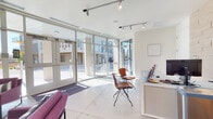 This single-floor leasing office presents a sleek, contemporary design that makes a strong first impression. The entrance is defined by expansive floor-to-ceiling glass windows and doors that flood the interior with abundant natural light while offering clear views of the surrounding urban streetscape. Polished concrete floors extend throughout, reinforcing the industrial-modern aesthetic.

The lobby features a comfortable lounge area anchored by a set of vibrant magenta upholstered chairs and a sofa, arranged around a round marble-top coffee table. A striking exposed concrete accent wall serves as a dramatic backdrop, adorned with a bold blue-and-white abstract artwork. Tall tropical plants add a touch of organic warmth to the otherwise clean, minimal palette. Track lighting overhead provides additional illumination and highlights the architectural details.

The leasing desk area transitions seamlessly from the lounge, featuring a light-toned stone tile accent wall and warm wood cabinetry beneath a wall-mounted flat-screen display. The service counter is equipped with a workstation, a stocked beverage mini-fridge, snack offerings, and informational materials, reflecting a hospitality-forward approach to the resident experience. The overall ambiance is bright, open, and professionally curated, blending industrial elements with modern comfort.