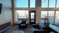 Located on Larson Lane West in University Place, Washington, this contemporary communal space is defined by its striking architectural design and an abundance of natural light. Soaring double-height ceilings create an open, airy atmosphere, while oversized windows arranged in dramatic tiers flood the interior with daylight and frame sweeping views of the surrounding landscape and sky.

The space is thoughtfully organized into two distinct zones. A comfortable lounge area features sleek black leather seating — including a sofa and armchairs — arranged around a mounted flat-screen television and a mid-century modern media console. Direct access to an outdoor terrace is provided through a glass-paneled door, offering an extension of the interior living space with views of the neighboring rooftop and treetops beyond.

The adjoining gathering and dining area is furnished with warm wood high-top tables and modern black bar-height chairs, ideal for collaborative work or casual socializing. Pendant linear lighting and industrial-style wall sconces cast a warm, focused glow over the tables, complemented by curated abstract artwork displayed on the soft gray walls. Patterned carpet tiles add texture throughout, while tiled flooring near a small service counter provides a practical transition zone. The overall ambiance is modern, polished, and inviting.