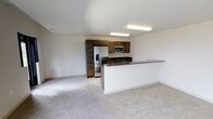 This single-floor space offers a clean, modern design centered around an open-concept layout that effortlessly connects the kitchen and living areas. The kitchen is well-appointed with rich dark wood cabinetry that provides generous storage space, complemented by dark granite countertops that add a touch of elegance. Stainless steel appliances — including a French door refrigerator with an ice and water dispenser, a built-in microwave, and a range — lend a sleek, contemporary feel. A peninsula-style breakfast bar extends from the kitchen, creating a natural transition into the adjacent living space and offering additional seating or prep space. The living area features plush, neutral-toned carpeting that adds warmth and comfort underfoot, while the light-toned tile flooring in the kitchen provides a practical and stylish contrast. The space is bathed in a bright, airy atmosphere thanks to white walls and flush-mounted fluorescent lighting overhead. Subtle architectural details, including clean baseboards and recessed ventilation, contribute to the overall polished and well-maintained character of the space. The open floor plan creates a spacious, cohesive environment that is both functional and inviting.