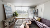 This single-floor residence blends industrial-inspired design with modern comfort throughout its well-appointed layout. The living area makes an immediate impression with expansive floor-to-ceiling windows that flood the space with natural light, casting dramatic patterns across the hardwood floors and textured area rug. Exposed piping along the ceiling adds an industrial edge, while whitewashed brick walls contribute warmth and character. A sectional sofa anchors the room alongside round wooden coffee tables and a lean ladder shelf, creating a relaxed yet stylish atmosphere. A secondary sitting area offers a more intimate setting with a sleek sofa, compact coffee table, and subtle ambient lighting.

The open kitchen features stainless steel appliances, including a range, over-the-range microwave, and French door refrigerator. Two-tone cabinetry—white uppers paired with warm wood lowers—frames a subway tile backsplash, while a peninsula with a white quartz countertop and bar seating serves as both a prep space and gathering spot beneath industrial pendant lighting.

The bedroom is a standout, offering panoramic floor-to-ceiling views of a waterfront landscape dotted with palm trees. Plush carpeting and abundant natural light enhance its serene ambiance. The bathroom presents a double-sink vanity with granite countertops, rich wood cabinetry, and hardwood flooring, combining functionality with refined style.