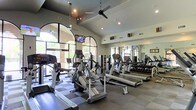 This single-floor fitness center offers a thoughtfully designed workout environment combining functionality with an open, airy atmosphere. The space is anchored by striking arched windows that line the walls, flooding the interior with abundant natural light and providing views of the surrounding outdoor greenery. Vaulted ceilings enhance the sense of spaciousness, while ceiling fans and recessed lighting ensure comfort throughout.

The cardio zone is well-stocked with multiple treadmills, elliptical trainers, and a stair climber, all arranged to allow ample space between equipment. A wall-mounted television is positioned for easy viewing during workouts. The flooring features a durable checkered tile pattern in the cardio area, providing stability underfoot.

A full-length mirror spans one wall, a practical addition that helps users monitor their form during exercise. Adjacent to the cardio equipment, a dedicated free weights section includes a neatly organized dumbbell rack. The overall ambiance is bright, clean, and professional, creating an inviting environment suited to a wide range of fitness routines.