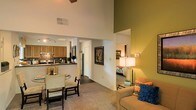 This single-floor residence offers a well-designed open-concept layout that seamlessly connects the primary living areas, creating a sense of spaciousness and flow throughout. The soaring vaulted ceilings are an immediate standout feature, dramatically amplifying the sense of openness in the main living space, which is accented by warm, earthy tones including a distinctive olive-green accent wall.

The kitchen is fully equipped with a range, microwave, and refrigerator, complemented by wooden cabinetry, a tiled backsplash, and a dark countertop. Track lighting illuminates the kitchen workspace effectively, while the layout connects conveniently to the dining area, which features a glass-top table with seating.

The open living area is carpeted and comfortably furnished, with a floor lamp providing warm ambient lighting and colorful accent cushions adding a welcoming touch. A bedroom is visible through an open corridor, offering a sense of privacy while remaining integrated with the overall layout. The hallway also provides access to additional rooms, including what appears to be a full bathroom. Artwork on the walls and thoughtful decor throughout contribute to an inviting, lived-in atmosphere suitable for comfortable everyday living.