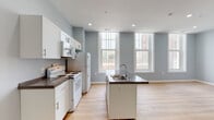 This single-floor unit features a clean, modern design characterized by soft blue-grey walls, light wood-tone flooring, and white trim throughout, creating a cohesive and airy aesthetic. Recessed lighting is found across all areas, ensuring consistent illumination throughout the space.

The kitchen is a standout feature, equipped with white cabinetry, dark countertops, and a central island with a built-in sink. White appliances include a range, refrigerator, and over-the-range microwave. Tall, multi-pane windows flood the kitchen and adjacent living area with abundant natural light and offer views of the surrounding urban landscape. The open layout connects the kitchen seamlessly to a generously sized living area, which also houses a dedicated in-unit laundry closet with a stacked washer and dryer.

A hallway connects the main living spaces to the private areas of the unit, including a well-proportioned bedroom featuring a tall, multi-pane window that continues the natural light theme found throughout. The bathroom presents a modern, streamlined vanity with a vessel-style sink, a large mirror with overhead lighting, and matching light wood flooring.