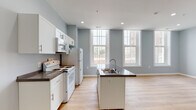 This single-floor unit features a clean, modern design characterized by soft blue-grey walls, light wood-tone flooring, and white trim throughout, creating a cohesive and airy aesthetic. Recessed lighting is found across all areas, ensuring consistent illumination throughout the space.

The kitchen is a standout feature, equipped with white cabinetry, dark countertops, and a central island with a built-in sink. White appliances include a range, refrigerator, and over-the-range microwave. Tall, multi-pane windows flood the kitchen and adjacent living area with abundant natural light and offer views of the surrounding urban landscape. The open layout connects the kitchen seamlessly to a generously sized living area, which also houses a dedicated in-unit laundry closet with a stacked washer and dryer.

A hallway connects the main living spaces to the private areas of the unit, including a well-proportioned bedroom featuring a tall, multi-pane window that continues the natural light theme found throughout. The bathroom presents a modern, streamlined vanity with a vessel-style sink, a large mirror with overhead lighting, and matching light wood flooring.