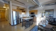 This single-floor urban loft showcases a seamless blend of industrial and contemporary design. The open-plan layout connects the kitchen and living areas, creating a spacious and cohesive environment. Exposed silver ductwork runs prominently across the ceiling, complemented by track lighting that adds both function and an industrial aesthetic to the space.

The well-appointed kitchen features warm honey-toned wood cabinetry, stainless steel appliances including a full-size refrigerator and dishwasher, a built-in microwave, and an electric range. A white subway tile backsplash adds a clean, classic contrast to the cabinetry. A built-in wine rack above the upper cabinets offers a convenient storage detail for entertaining.

At the center of the kitchen stands a movable island with a white surface and industrial-style metal bar stools, serving as both a prep area and a casual dining spot. Polished concrete floors extend throughout, reinforcing the loft's industrial character while remaining easy to maintain.

The living area transitions naturally from the kitchen, offering room for comfortable seating and décor. Natural light filters into the space, enhancing the open and airy feel. Overall, this loft delivers a functional, stylish environment with distinctive architectural character.
