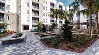 Located at 3600 E Fletcher Ave in Tampa, Florida, this outdoor courtyard space serves as a central gathering area within a contemporary multi-story residential complex. The architecture blends modern white stucco facades with warm brick detailing, accented by bold cobalt blue entry doors and railings that add a distinctive visual identity to the development.

The courtyard is thoughtfully designed with a network of patterned paver walkways that connect multiple seating areas and building entrances. Lush tropical landscaping is a defining feature, with tall, mature palm trees, ferns, shrubs, and layered plantings set within mulched garden beds. The greenery creates a welcoming, resort-like atmosphere that reflects the Florida climate beautifully.

Several seating nodes are distributed throughout the space, including vibrant, colorful lounge chairs and outdoor sofas in aqua and orange tones, as well as a central fire pit or water feature surrounded by bench seating. String lights are strung above portions of the courtyard, adding a warm ambiance for evening use. A pergola structure anchors one end of the space, providing shaded seating. The open-air design allows for abundant natural light throughout the day, making this courtyard a lively and functional outdoor amenity for the surrounding community.