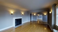 This single-floor space offers an open, flowing layout characterized by warm hardwood flooring that extends throughout the main living areas. The living room serves as a focal point, anchored by a classic fireplace with a crisp white surround and striking dark marble surround and hearth, adding an elegant contrast to the neutral palette. Soft gray walls create a calm, cohesive backdrop, while wall-mounted sconces provide warm ambient lighting throughout the space. Arched doorways connect the living area to additional rooms, lending architectural interest and a sense of continuity. One arch leads to a secondary living or dining area, and another provides a view into what appears to be a kitchen, featuring light cabinetry visible in the background. The generous proportions of the main living area allow for versatile furniture arrangements. The combination of refined architectural details — including clean white baseboards, smooth ceilings, and the arched passageways — give the space a timeless, understated character well-suited to a variety of interior styles.
