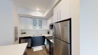 This single-floor residence showcases a thoughtfully designed interior with a strong emphasis on modern aesthetics and functional living. The kitchen stands as a centerpiece of the space, featuring a striking two-tone design with crisp white upper cabinetry and dark lower cabinets that create a bold, contemporary contrast. White quartz countertops provide ample prep space and complement the clean lines of the cabinetry. Stainless steel appliances, including a full-size refrigerator, built-in microwave, and electric range, lend a polished, professional feel to the cooking space. Glass-front upper cabinets add a touch of elegance while offering display storage. Recessed lighting illuminates the kitchen from above, highlighting the tray ceiling detail that adds architectural interest. The warm wood-look flooring grounds the space and flows seamlessly throughout, offering a natural contrast to the modern cabinetry. A single-basin sink is neatly integrated into the countertop beneath a sleek faucet. Overall, the space presents a clean, well-appointed interior that balances practicality with contemporary style, making it an inviting and efficient place to live.