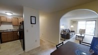 This 2-bedroom, 1.5-bathroom single-floor residence offers a cohesive and functional layout designed for comfortable everyday living. The interior is finished with neutral cream-toned walls and wall-to-wall carpeting, creating a warm and inviting atmosphere throughout. A defining architectural detail is the elegant arched passageway that connects the dining area to the living room, adding a classic touch to the open floor plan. The living room is a spacious and well-lit area, accommodating a full sofa set and accented by floor lamps that contribute to a cozy ambiance. A window allows natural light to filter into the space. The dining area, visible in the foreground, provides ample room for a full dining table setup, making it well-suited for everyday meals or entertaining. The kitchen is accessible nearby, with dark cabinetry offering practical storage. The hallway connects the various zones of the unit seamlessly, maintaining an easy flow between spaces. With two bedrooms and one and a half bathrooms, this residence offers a practical and comfortable layout suitable for a range of lifestyles.