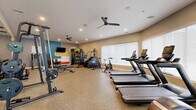 Located at 3755 E McKinney St in Denton, Texas, this fitness center offers a comprehensive exercise facility within a single, thoughtfully designed room. The space features speckled, durable flooring throughout, providing a clean and functional foundation suited for high-traffic use. Large windows along the perimeter flood the room with natural light and offer views of the surrounding outdoor environment, while ceiling fans and recessed lighting ensure year-round comfort and visibility.

The cardio zone is well-appointed with multiple treadmills, elliptical machines, stationary bikes, a stair climber, and a rowing machine, providing a wide range of aerobic options. The strength training area includes a multi-functional cable machine, adjustable weight benches, and a full rack of dumbbells neatly organized on tiered storage racks. Full-length mirrors along one wall enhance the sense of space and allow users to monitor their form during workouts.

A mounted flat-screen television and a colorful accent wall featuring teal, yellow, and red geometric shapes add visual energy to the space. The entrance features a glass-panel door that connects to the broader property. Ceiling fans are strategically placed throughout to maintain airflow, contributing to a comfortable workout environment. Overall, this fitness center is well-organized, modern, and fully equipped to support a diverse range of exercise routines.