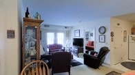 This single-floor residence offers a warm and inviting atmosphere with a traditionally styled interior. The living area serves as the heart of the space, featuring a comfortable seating arrangement that includes upholstered armchairs, a loveseat, and a leather recliner positioned around a central area rug. A dark wood media console houses a flat-screen television, making it a functional entertainment hub. Natural light filters into the room through a set of French doors dressed with white curtains, brightening the neutral-toned walls and light carpeted flooring. A classic oak display cabinet stands prominently in one corner, showcasing a curated collection of decorative glassware and keepsakes. Framed artwork and a decorative wall clock add personal character throughout the space. An open doorway leads to an adjacent sitting room, which is visible from the main living area and features a red sofa, reinforcing the cohesive and homey aesthetic. The layout encourages a natural flow between living spaces, making the home feel connected and well-organized. Overall, the space combines functional design with a classic, lived-in charm suited for comfortable everyday living.
