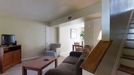 This two-story residence offers a thoughtfully arranged layout that balances comfort and functionality. The main living area is furnished with a plush sofa, an upholstered armchair, and a wooden coffee table, creating an inviting space for relaxation. Neutral-toned walls and light carpeting throughout lend a calm, understated aesthetic to the interior. Ceiling vents are integrated into the design, ensuring efficient climate control across the space. A staircase with a warm wooden banister connects the two floors, adding a classic architectural touch. Adjacent to the living area, the dining space accommodates a wooden table and chairs, making it well-suited for everyday meals and gatherings. Framed wall art and touches of greenery add character to the dining area. The open sightlines between the living and dining areas create a sense of continuity and spaciousness. Overall, the residence presents a clean, practical interior with a neutral palette, providing a versatile backdrop that can be easily personalized to suit a variety of styles and preferences.