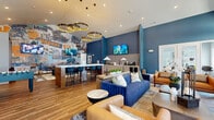 This expansive single-floor amenity space is thoughtfully designed to offer a dynamic mix of recreation, relaxation, and productivity. The main lounge area makes an immediate impression with soaring ceilings, bold blue accent walls, and a large-scale Milwaukee-themed mural that serves as a striking focal point. Stacked circular gold pendant lights illuminate a central island with bar seating, while a dedicated beverage station and two fully equipped kitchens—featuring stainless steel appliances, shaker-style cabinetry with gold hardware, and mosaic tile backsplashes—provide excellent functionality for gatherings. A separate living area showcases a stone-surround linear fireplace, a wall-mounted TV, rich hardwood flooring, and curated furnishings including a tufted leather sofa and swivel armchairs. The game room extends the entertainment offerings with a billiard table, foosball table, and cozy lounge alcoves with pendant lighting. The fitness center is well-appointed with strength and cardio equipment, patterned hardwood floors, and dual ceiling fans. A private office with patterned wallpaper, natural light, and a glass-panel door provides a quiet workspace. The restroom features wood-look flooring, a textured accent wall, and ADA-accessible fixtures. Three connecting hallways ensure seamless flow throughout.