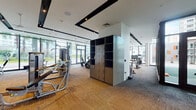 This contemporary fitness center is designed to deliver a comprehensive workout experience within a bright, open, and thoughtfully organized single-floor layout. Large floor-to-ceiling windows and glass doors line multiple walls, flooding the space with natural light and offering views of lush palm trees and the surrounding landscape, creating an energizing atmosphere throughout.

The strength training area is outfitted with a full suite of professional-grade Cybex Prestige machines, including cable systems, seated press units, and various resistance stations, all arranged to allow ample circulation. Full-length mirrors amplify the sense of space and support proper form during training sessions. A central locker unit provides convenient storage for personal belongings.

The dedicated cardio zone features multiple Cybex treadmills, elliptical trainers, and stationary bikes, positioned to face large windows and wall-mounted flat-screen televisions — ensuring an engaging workout experience. The flooring transitions between textured carpet tiles and rubberized matting, delineating functional zones while providing comfort and durability underfoot.

A glass-enclosed entryway leads to an outdoor training area visible through wide double doors, extending the fitness options beyond the interior. The white ceiling with dark accent beams and recessed track lighting adds a sleek, modern aesthetic, making this fitness center as visually refined as it is functionally equipped.