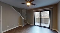 This two-story residence is thoughtfully designed with a functional layout that separates living and sleeping areas across its two levels. The main level features wood-style flooring throughout, lending a clean, cohesive look to the living and dining spaces. A ceiling fan with a built-in light fixture anchors the living area, which is filled with natural light from a large sliding glass door that opens to a private balcony overlooking mature trees. The kitchen is accessible via a pass-through opening, offering a practical connection between the cooking and dining areas. The kitchen is equipped with stainless steel appliances, dark cabinetry, and a granite-style countertop that extends to the pass-through bar. A utility area with an electrical panel and wall-mounted connections is also present on this level. The staircase, accented by a white handrail, leads to the upper level, where carpeted bedrooms provide a comfortable retreat. The upper landing features an open railing overlooking the stairwell, with multiple doors leading to the bedrooms. The bathroom includes a dark wood vanity, stone-patterned countertop, framed mirror, and tiled shower surround, offering a polished and well-appointed finish.