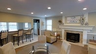 This single-floor space offers a thoughtfully designed open-concept layout that seamlessly connects the main living, dining, and kitchen areas. The living room is anchored by a classic fireplace with a white decorative mantel, creating a warm and inviting focal point. Neutral-toned walls and recessed ceiling lighting throughout lend a clean, comfortable ambiance. Plush accent chairs and a glass-top dining table furnish the space with a balance of style and practicality.

A pass-through window connects the living and dining area to the kitchen, which features light tile flooring, a raised breakfast bar with bar stools, and full-size appliances including a refrigerator. The kitchen provides ample functionality while maintaining visual openness to the rest of the space.

Carpeted flooring in the living area transitions smoothly to tile in the kitchen and dining zone, defining each space while maintaining a cohesive flow. Natural light filters in through double-hung windows dressed with blinds, complementing the recessed lighting above. The overall design emphasizes comfort and practicality, making this a versatile and welcoming single-level living space.