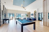 Located at 112 Point Pl in Mooresville, NC, this thoughtfully designed single-floor space serves as a vibrant community lounge and workspace. The interior blends coastal-inspired aesthetics with modern sophistication through a cohesive palette of teal, warm gold, and crisp white tones throughout.

The living area is the heart of the space, boasting soaring ceilings that amplify its openness. Large floor-to-ceiling windows flood the room with natural light and offer views of an outdoor pool area. Rich light oak hardwood flooring runs throughout, complementing plush blue velvet seating arranged around a marble-topped coffee table. A striking textured wave-panel accent wall in teal frames a dining nook outfitted with tan leather booth seating, dark bistro tables, and elegant gold pendant lighting.

The two office spaces offer a more focused atmosphere. Each features a sleek marble-surface desk, a cognac leather chair, and refined geometric patterned wallcovering, creating a calm and productive environment. Soft overhead pendant lighting and lush greenery add warmth to each workstation.

Curated artwork, including a bold grid of abstract panels, adds a gallery-like character to the lounge. The seamless blend of functional zones and design-forward details makes this space both practical and visually compelling.