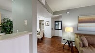 This single-floor residence offers a thoughtfully designed layout that balances comfort and functionality throughout its living spaces. Rich, dark hardwood-style flooring flows through the main living and dining areas, creating a warm and cohesive aesthetic.

The living room is a welcoming focal point, featuring a sliding glass door framed by vertical blinds that fills the space with natural light and provides a view of the greenery outside. The adjacent dining area is defined by a partial wall partition, lending a sense of structure while maintaining an open feel. A built-in shelving nook adds convenient storage and display space nearby.

The galley-style kitchen is efficiently organized with light wood cabinetry lining both walls, offering generous counter and storage space. Black appliances, including a gas range, microwave, dishwasher, and refrigerator, provide a functional contrast to the light cabinetry and neutral countertops.

The two bedrooms are carpeted for comfort and each feature ceiling fans with light fixtures. The primary bedroom includes access to a walk-in closet and an adjoining bathroom. The bathroom is clean and practical, with a single-sink vanity topped with light countertops, a large mirror, and light-up vanity fixtures above.

Soft, neutral tones in sage green and warm gray are carried throughout the walls, contributing to a calm and cohesive ambiance across the entire residence.