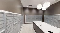This single-floor mail room is designed with both functionality and aesthetics in mind. The space is lined on multiple walls with built-in banks of individual mailboxes finished in brushed aluminum, offering ample mail storage capacity for a large number of occupants. The mailbox units are framed in crisp white trim that contrasts cleanly against the warm taupe-toned walls, lending the room a polished, contemporary feel. At the center of the room stands a dark wood-toned sorting island topped with a light-colored surface, providing a convenient workspace for sorting and organizing deliveries. Two circular drop-in openings on the island surface add a practical touch for mail handling. The flooring is a light-toned wood-look material that enhances the room's brightness and modern character. Overhead, a combination of recessed lighting and two striking globe-style pendant lights with an intricate, layered design illuminate the space evenly while serving as subtle decorative focal points. The overall ambiance is clean, organized, and professionally finished, reflecting a thoughtfully designed communal service space.
