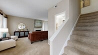 This two-story residence offers a thoughtfully designed layout with neutral tones and plush carpeting throughout the main living areas. The living room serves as a central gathering space, anchored by a white brick fireplace and framed by multiple windows with floor-length curtains that invite natural light. A staircase with white-painted railings connects to the upper level, accompanied by a convenient pass-through window detail.

The kitchen features white cabinetry, grey countertops, and wood-look vinyl flooring, offering a clean and functional workspace equipped with a dishwasher and ample storage. Adjacent to the kitchen, the dining area benefits from a large window that fills the space with natural light, creating an inviting atmosphere.

The primary bedroom is a standout space, boasting soaring vaulted ceilings, a ceiling fan, soft carpeting, built-in shelving, and a sliding glass door leading to an outdoor area. A spacious en-suite bathroom includes a dual-sink vanity with grey countertops. A second bedroom offers a comfortable, well-proportioned space with warm, neutral tones. A full bathroom with wood-look flooring and a tub-shower combination completes the layout.