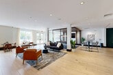 Located at 1000 Ellis Drive in Glen Mills, Pennsylvania, this thoughtfully designed amenity space offers a generously proportioned, single-floor layout ideal for socializing and relaxation. Light-toned hardwood-style flooring flows continuously throughout, unifying the various zones and lending a warm, contemporary aesthetic.

The open lounge area serves as the heart of the space, furnished with a plush sofa, leather accent chairs, and a sleek coffee table arranged atop a patterned area rug. Recessed lighting illuminates the white ceilings, while large windows invite natural light and views of the surrounding greenery. A decorative metal screen partition adds architectural interest, subtly defining zones without disrupting the open flow.

A dedicated billiards area features a full-sized pool table beneath a wood-paneled ceiling detail and curated wall art, creating an inviting game room atmosphere. Adjacent to this is a quiet lounge nook accented by a deep teal feature wall, comfortable armchairs, and glass-walled enclosures that provide privacy while maintaining visual connectivity.

A dining banquette area offers built-in seating with marble-topped tables and warm wood chairs beside large windows, perfect for casual gatherings. A fully equipped kitchenette with dark wood cabinetry and bar seating rounds out the amenities. A glass-enclosed staircase visible from the lounge adds a striking architectural focal point to the overall design.