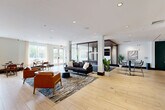 Located at 1000 Ellis Drive in Glen Mills, Pennsylvania, this thoughtfully designed amenity space offers a generously proportioned, single-floor layout ideal for socializing and relaxation. Light-toned hardwood-style flooring flows continuously throughout, unifying the various zones and lending a warm, contemporary aesthetic.

The open lounge area serves as the heart of the space, furnished with a plush sofa, leather accent chairs, and a sleek coffee table arranged atop a patterned area rug. Recessed lighting illuminates the white ceilings, while large windows invite natural light and views of the surrounding greenery. A decorative metal screen partition adds architectural interest, subtly defining zones without disrupting the open flow.

A dedicated billiards area features a full-sized pool table beneath a wood-paneled ceiling detail and curated wall art, creating an inviting game room atmosphere. Adjacent to this is a quiet lounge nook accented by a deep teal feature wall, comfortable armchairs, and glass-walled enclosures that provide privacy while maintaining visual connectivity.

A dining banquette area offers built-in seating with marble-topped tables and warm wood chairs beside large windows, perfect for casual gatherings. A fully equipped kitchenette with dark wood cabinetry and bar seating rounds out the amenities. A glass-enclosed staircase visible from the lounge adds a striking architectural focal point to the overall design.