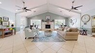 Located at 98 Paddock Club Dr in Panama City Beach, Florida, this single-floor space seamlessly blends a living room and dining area into one welcoming, open-plan layout. The interior is defined by striking vaulted ceilings with exposed beam detailing, anchored by dual ceiling fans that reinforce the airy, coastal atmosphere. Light-toned tile flooring runs throughout the main area, transitioning to warm hardwood in the hallway corridor, adding visual variety and texture.

The living room is a spacious, well-lit gathering area centered around a classic fireplace with a white surround and decorative mantle. A plush neutral sofa, accent armchairs, and a soft area rug in coastal blue tones create an inviting seating arrangement. An abundance of large windows along the rear wall floods the space with natural light and offers direct views of the outdoor pool area.

The dining area features a round glass-top table with wicker chairs, positioned beside French doors and expansive windows that further enhance the connection to the outdoors. Soft sage-green walls with white wainscoting paneling lend the entire space a refined yet relaxed coastal character. The hallway connects to additional rooms via elegant French doors with glass pane inserts, maintaining openness and light flow throughout.