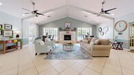 Located at 98 Paddock Club Dr in Panama City Beach, Florida, this single-floor space seamlessly blends a living room and dining area into one welcoming, open-plan layout. The interior is defined by striking vaulted ceilings with exposed beam detailing, anchored by dual ceiling fans that reinforce the airy, coastal atmosphere. Light-toned tile flooring runs throughout the main area, transitioning to warm hardwood in the hallway corridor, adding visual variety and texture.

The living room is a spacious, well-lit gathering area centered around a classic fireplace with a white surround and decorative mantle. A plush neutral sofa, accent armchairs, and a soft area rug in coastal blue tones create an inviting seating arrangement. An abundance of large windows along the rear wall floods the space with natural light and offers direct views of the outdoor pool area.

The dining area features a round glass-top table with wicker chairs, positioned beside French doors and expansive windows that further enhance the connection to the outdoors. Soft sage-green walls with white wainscoting paneling lend the entire space a refined yet relaxed coastal character. The hallway connects to additional rooms via elegant French doors with glass pane inserts, maintaining openness and light flow throughout.