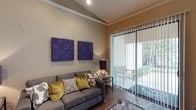 This single-floor space showcases a thoughtfully designed living area that blends comfort with contemporary style. The living room draws immediate attention with its vaulted ceiling adorned with elegant crown molding, adding a sense of architectural character and height to the room. Warm, neutral-toned walls provide a versatile backdrop that complements the bold purple wall art panels, which serve as striking focal points above the plush gray sectional sofa. The sofa is dressed with an eclectic mix of accent pillows in gold, ivory, and charcoal, lending warmth and personality to the space. Rich hardwood flooring runs throughout, grounding the room with a sense of warmth and continuity. A glass-top coffee table and a side table with a deep burgundy lamp complete the seating arrangement. Natural light floods the room through a large sliding glass door fitted with vertical blinds, offering both privacy and easy control of light. The door opens directly to a private outdoor patio area, where lush greenery is visible, seamlessly connecting the interior to the outdoors. The overall ambiance is sophisticated yet inviting, making this a well-appointed space suited for both relaxation and entertaining.