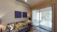 This single-floor space showcases a thoughtfully designed living area that blends comfort with contemporary style. The living room draws immediate attention with its vaulted ceiling adorned with elegant crown molding, adding a sense of architectural character and height to the room. Warm, neutral-toned walls provide a versatile backdrop that complements the bold purple wall art panels, which serve as striking focal points above the plush gray sectional sofa. The sofa is dressed with an eclectic mix of accent pillows in gold, ivory, and charcoal, lending warmth and personality to the space. Rich hardwood flooring runs throughout, grounding the room with a sense of warmth and continuity. A glass-top coffee table and a side table with a deep burgundy lamp complete the seating arrangement. Natural light floods the room through a large sliding glass door fitted with vertical blinds, offering both privacy and easy control of light. The door opens directly to a private outdoor patio area, where lush greenery is visible, seamlessly connecting the interior to the outdoors. The overall ambiance is sophisticated yet inviting, making this a well-appointed space suited for both relaxation and entertaining.
