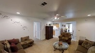 This single-floor home offers a welcoming and functional layout with a warm, lived-in character. The living room serves as the central gathering space, featuring neutral carpeted flooring, white walls accented with decorative wall art, and a ceiling fan with integrated lighting that enhances comfort and airflow. The room is generously sized, accommodating plush upholstered armchairs and a glass-top coffee table with an elegant wrought-iron base, creating a cozy yet practical seating arrangement. A large dark wood armoire adds a classic touch and serves as a natural focal point. An open pass-through connects the living area to the kitchen, which features warm wood cabinetry, light countertops, and a raised breakfast bar with seating. The kitchen flows naturally into a dining area, illuminated by natural light from nearby windows dressed in draped curtains. The dining space comfortably accommodates a table and chairs, making it ideal for everyday meals. Throughout the home, the consistent use of neutral tones and soft materials creates a cohesive, approachable atmosphere. A doorway from the living room provides access to additional areas of the home, reinforcing the practical and connected flow of this single-level residence.